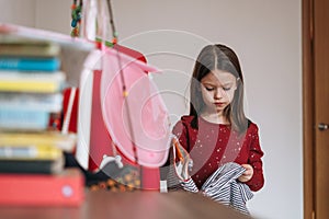 Thinking tween little girl with long dark hair in red dress among her beautiful dresses in wardrobe in children`s room at home
