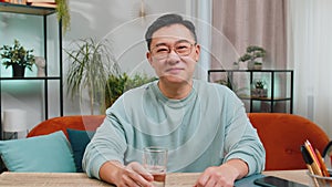 Thirsty Asian man sits at home table holding glass of natural aqua make sips drinking filtered water