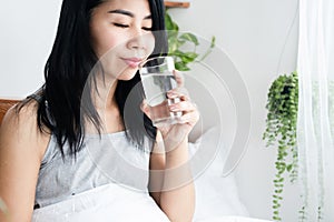 Thirsty Asian woman drinking fresh water from glass after wakeup in morning sitting in bed photo