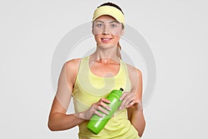 Thirsty beautiful active woman takes break after playing tennis, dressed in casual sportswear, holds bottle with water, stands alo