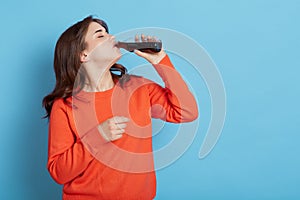 Thirsty female wearing orange jumper standing isolated over blue background  holding bottle of fizzy water and drinking  adorable