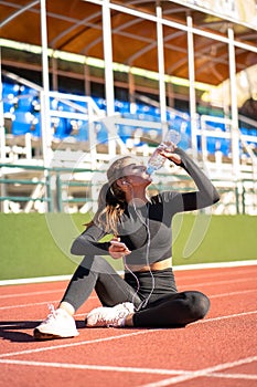 Thirsty fit woman drinking water from plastic bottle, resting after jogging on a treadmill rubber stadium on sunny summer day
