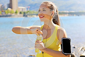 Thirsty fitness woman opens bottle of water after training outdoor. Fit woman using smartphone fitness app on armband as activity