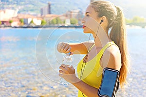 Thirsty fitness woman opens bottle of water after training outdoor. Fit woman using smartphone fitness app on armband for