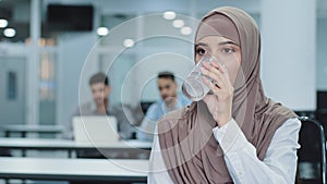 Thirsty millennial indian arabic female worker in hijab holding glass drinking pure mineral water in office, thoughtful