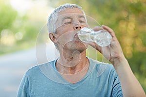 Thirsty senior male drinks water from palstic bottle, being tired after cardio training, has active lifestyle, dressed casually, p