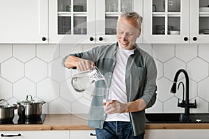 Thirsty Senior Man Pouring Water From Jug To Glass In Kitchen Interior