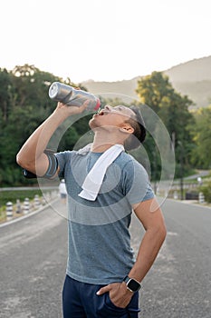 A thirsty, tired Asian man in sportswear is drinking water from a bottle after a long run outdoors
