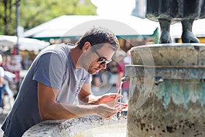 Thirsty young casual cucasian man drinking water from public city fountain on a hot summer day