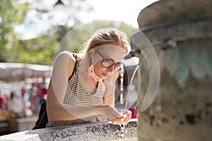 Thirsty young casual cucasian woman wearing medical face mask drinking water from public city fountain on a hot summer