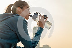 Thirsty young woman runner is resting and drinking water bottle with headphone after morning cardio on street outdoors in the city