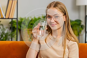 Thirsty young woman sitting at home holding glass of natural aqua make sips drinking still water