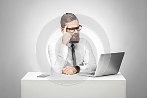 Thoughful bearded young boss in white shirt and black tie are sitting in office on desk and watching daily report on laptop,