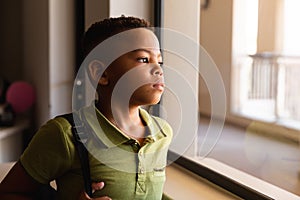 Thoughtful african american elementary schoolboy standing by window while standing in classroom