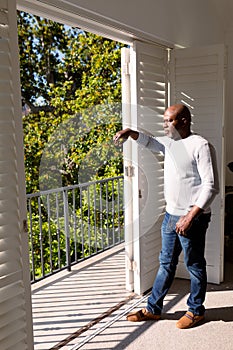 Thoughtful african american senior man standing at window and looking into distance