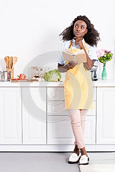 thoughtful african american woman in apron holding cookbook