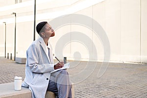 thoughtful african business woman sitting with a notebook and a pen on a bench outdoors