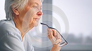 Thoughtful aged woman in blue blouse holds designed glasses in wrinkly hand near mouth against blurred window at home