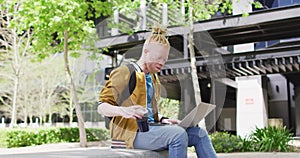 Thoughtful albino african american man with dreadlocks sitting in park drinking coffee, using laptop