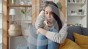Thoughtful Asia lady sit embracing knees at sofa in living room at house look outside with feel lonely, Sad depressed teenager