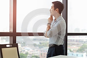 Thoughtful asian business man standing next to big window looking out.