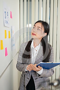 A thoughtful Asian businesswoman is looking at the ideas on sticky notes on the wall