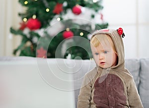 Thoughtful baby in deer suit near Christmas tree