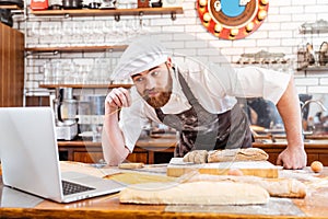 Thoughtful baker cutting bread and using laptop on kitchen