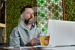 Thoughtful bearded man in casual clothes in cafe using laptop