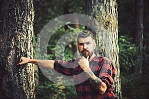 Thoughtful bearded man in lumberjack shirt with tattoo on his arm wandering in forest. Lone hiker exploring wonders of