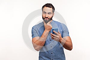 Thoughtful bearded man scratching chin holding smartphone in hand, contemplating about software updating, choosing suitable