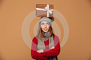 Thoughtful beautiful girl in winter clothes standing with crossed hands with gift box on her head, looking aside