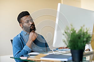 Thoughtful black businessman sitting at workplace and looking at computer screen, thinking about problem solution
