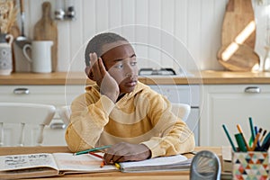 Thoughtful bored Black schoolboy look to side doing homework sits at table with textbooks at home