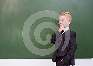 Thoughtful boy in a suit standing near empty green chalkboard