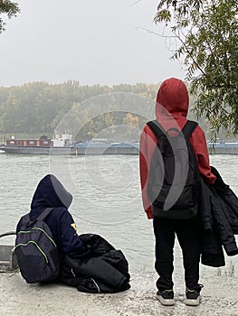 Thoughtful boys with hoods looking a cago ship sailing down Danube river. Cold and calm winter day concept.