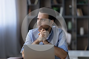 Thoughtful businessman sitting at desk with laptop, looking in distance