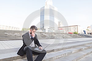 A thoughtful businessman sitting on a staircase with a laptop on his knees against a backdrop of urban landscape