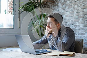 Thoughtful businessman touching chin, pondering ideas or strategy, sitting at wooden work desk with laptop, freelancer working on