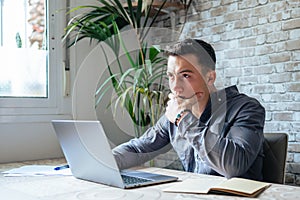 Thoughtful businessman touching chin, pondering ideas or strategy, sitting at wooden work desk with laptop, freelancer working on