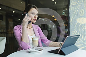 Thoughtful businesswoman talking on the phone with clients or business partners while looking at laptop monitor at cafe