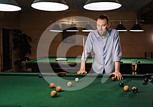 Thoughtful caucasian bearded man plays billiards, thinking and looking on pool. Billiard room on the background. Hobby