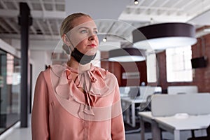 Thoughtful caucasian businesswoman wearing lowered face mask standing in office looking away