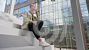 Thoughtful caucasian guy with a hearing aid sits on the stairs in the city center.