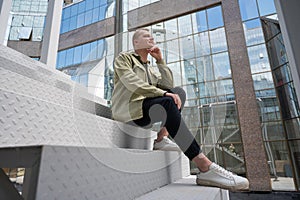 Thoughtful caucasian guy with a hearing aid sits on the stairs in the city center.