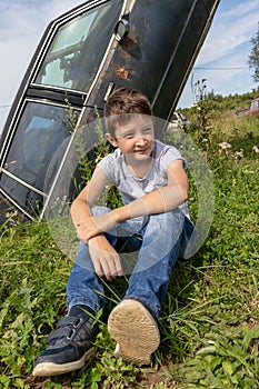 Thoughtful caucasian teenage boy sitting on the grass in countyside in summer day with serious face expression, a retro car is on