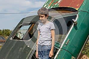 Thoughtful caucasian teenage standing on the grass in countyside in summer day with serious face expression, a retro car is on