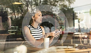 Thoughtful caucasian woman holding mobile phone while looking through the coffee shop window during coffee break.