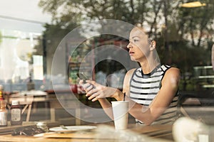 Thoughtful caucasian woman holding mobile phone while looking through the coffee shop window during coffee break.