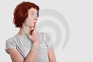 Thoughtful contemplative woman stands sideways, keeps index finger on lips, dressed in grey t shirt, contemplates about something